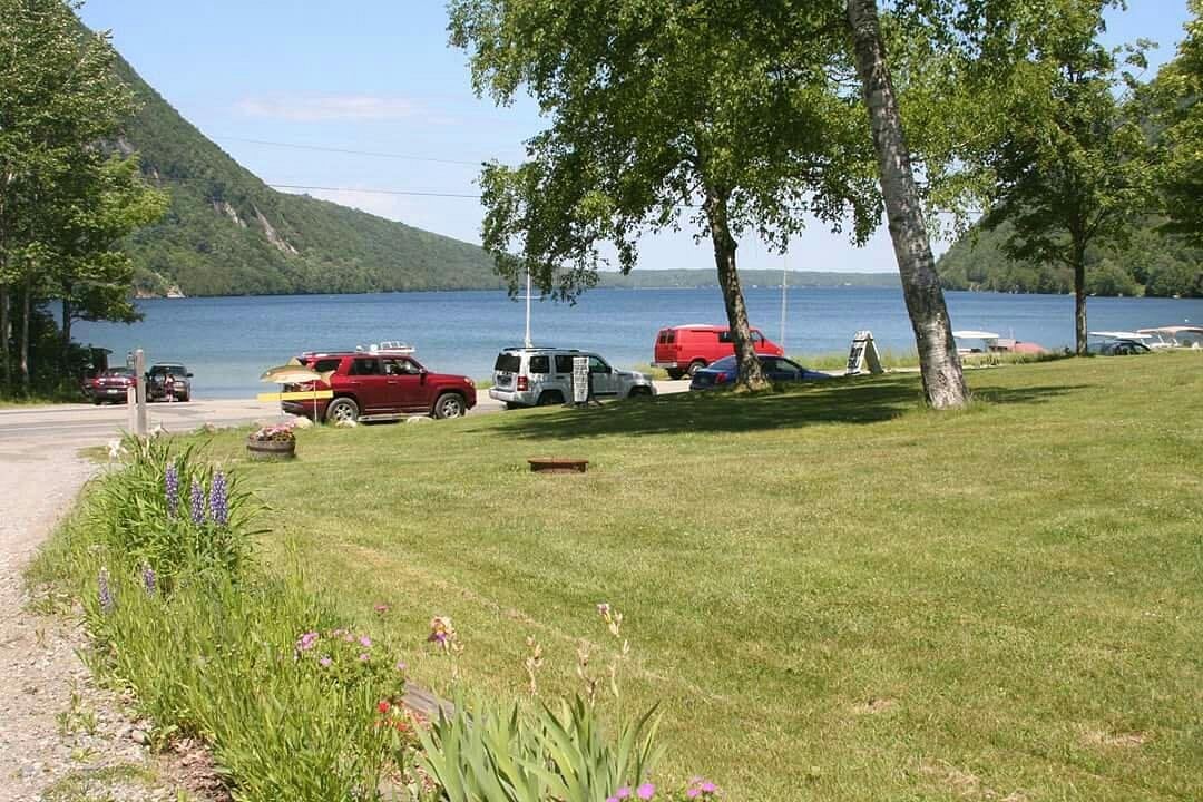 Lake Willoughby shoreline access with grass, trees, parked cars, and mountain views.